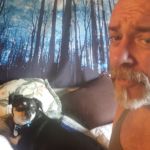 A man sitting on a bed with a tapestry of a forest in the evening with bare trees, and a dog laying on pillows looking at the man